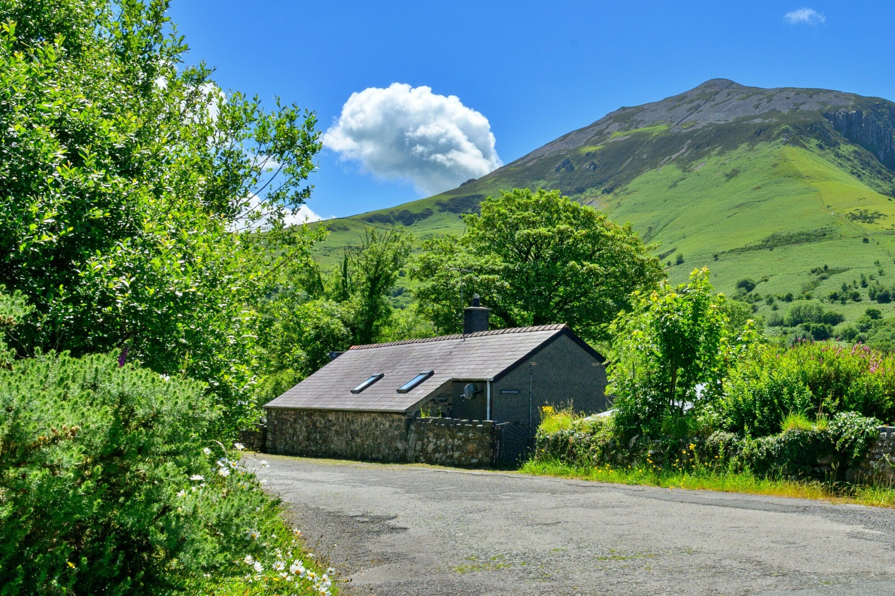 Une maison en pierre avec un toit en pente et deux lucarnes, entourée d’une végétation luxuriante et avec une montagne en arrière-plan.