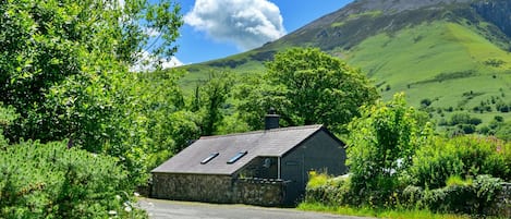 A stone house with a sloped roof and two skylights, surrounded by lush greenery and a mountain in the background.