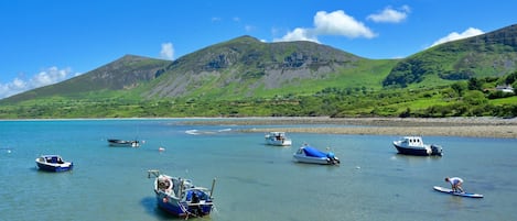 A coastal scene with boats on calm water, a clear sky, and green hills in the background.