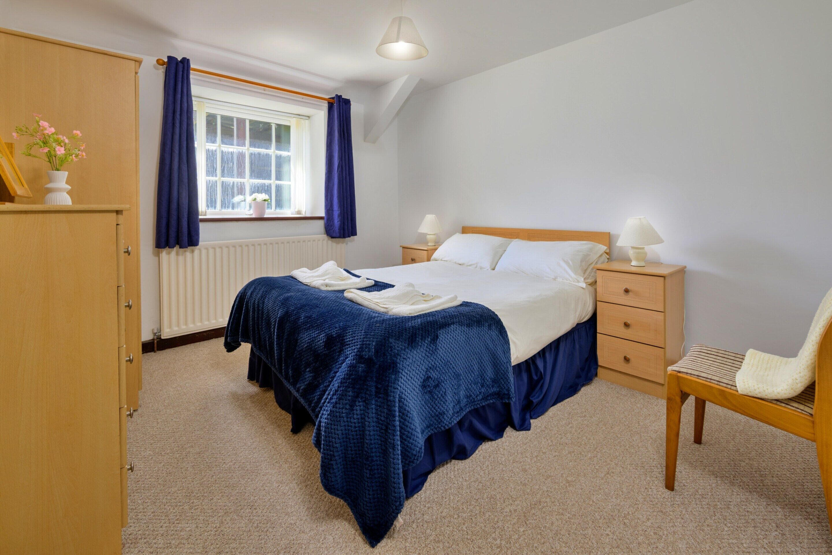 A bedroom with a bed, bedside tables, a chair, a window with curtains, a radiator, and a lamp.