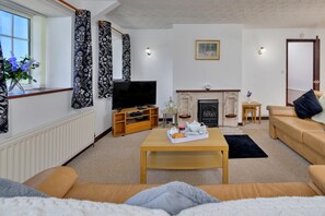 A living room with a sofa, a coffee table, a television, a fireplace, and a window with curtains. - Charming Dorset Coach House for 6 (Caernarfon)