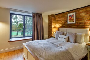 A bedroom with a large window, a brick wall, a bed with pillows, and a nightstand with a lamp. - Stylish Barn Retreat for 8 (Wimborne)
