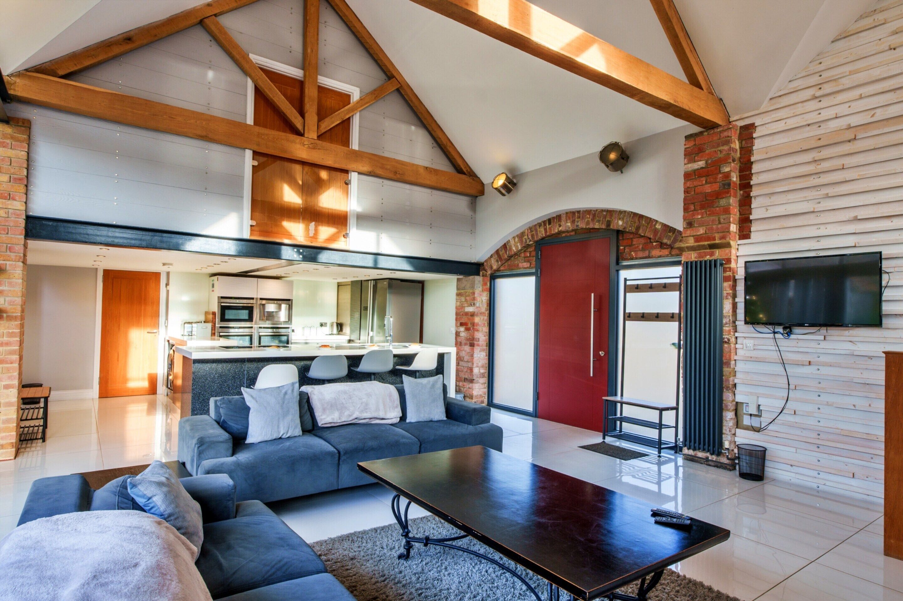 A spacious living area with a high ceiling, exposed wooden beams, a modern kitchen, and a red door.