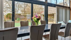 A dining area with a table and chairs, a vase of flowers, and a view of a garden through large windows. - Stylish Barn Retreat for 8 (Wimborne)