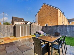 A patio with a glass-top table and wicker chairs, a wooden fence, and a brick building in the background. - House Near Golf Course Llanelli (Llanelli)