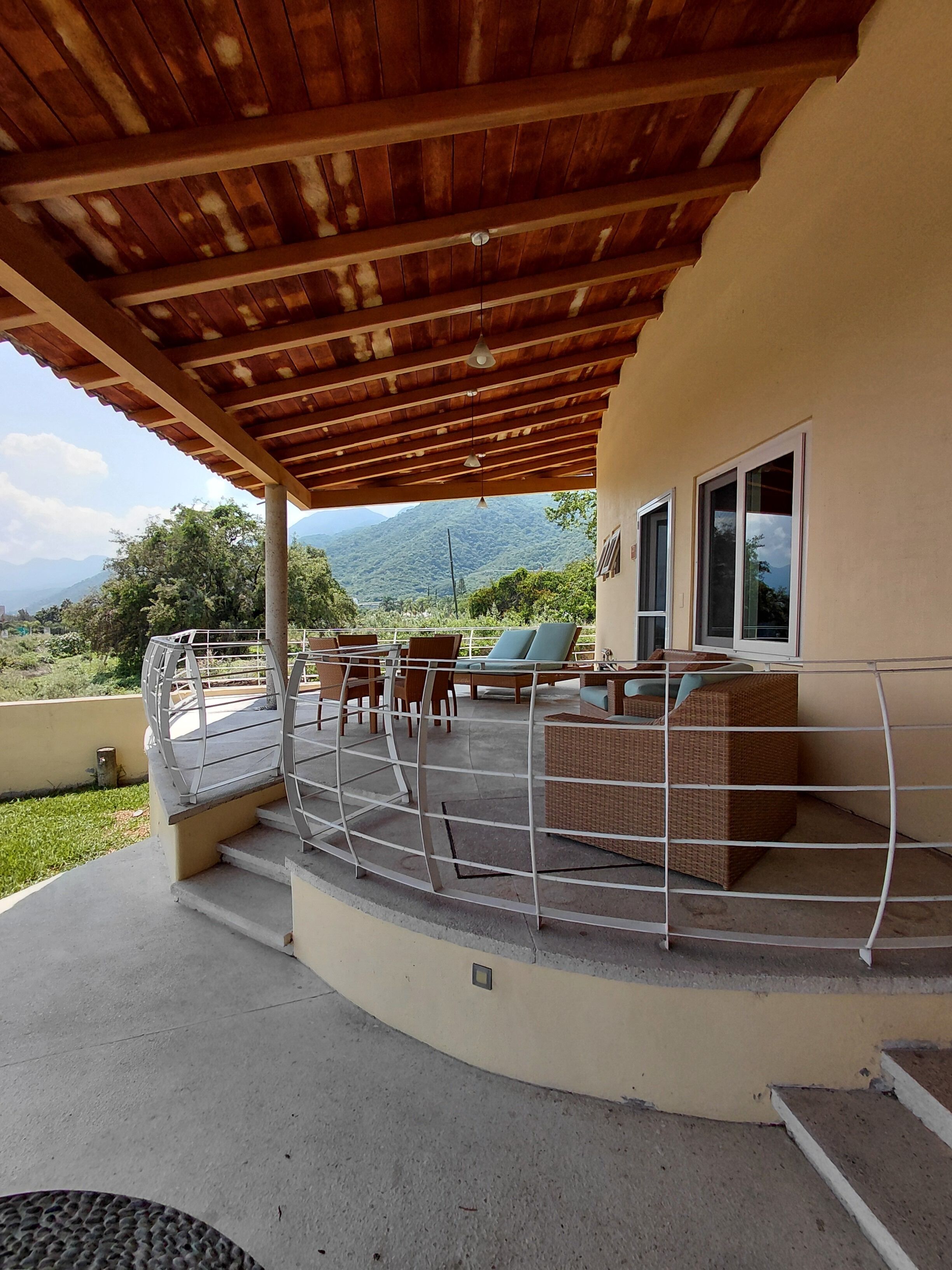 A covered outdoor seating area with a view of mountains, a railing, and a mix of seating options.