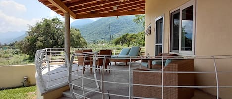 A covered outdoor seating area with a view of mountains, a railing, and a mix of seating options.
