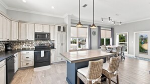 A modern kitchen with white cabinetry, a central island, and a dining area with wicker chairs. - Barefoot Treasure  Gulf Views, Canal Front & Pool (Navarre)