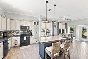 A modern kitchen with white cabinetry, a central island, and a dining area with wicker chairs. - Barefoot Treasure  Gulf Views, Canal Front & Pool (Navarre)