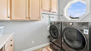 A laundry room with a washing machine and dryer, wooden cabinets, a laundry basket, and a window with a view of the sky. - Barefoot Treasure  Gulf Views, Canal Front & Pool (Navarre)