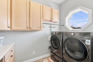 A laundry room with a washing machine and dryer, wooden cabinets, a laundry basket, and a window with a view of the sky. - Barefoot Treasure  Gulf Views, Canal Front & Pool (Navarre)