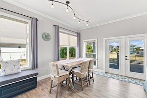 A dining area with a wooden table and wicker chairs, a clock on the wall, and a view of the outside through large windows. - Barefoot Treasure Gulf Views, Canal Front & Pool (Navarre)
