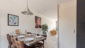 Ein Essbereich mit einem Holztisch, gedeckt für sechs Personen, umgeben von Stühlen. An der Wand befinden sich zwei gerahmte Kunstwerke, ein Topfpflanze und eine Tür zu einem anderen Raum.