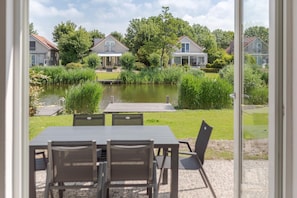 A view from a window of a dining area with a table and chairs, overlooking a pond with a wooden dock, surrounded by greenery and houses. - Waterfront Bungalow With Garden (Makkum)