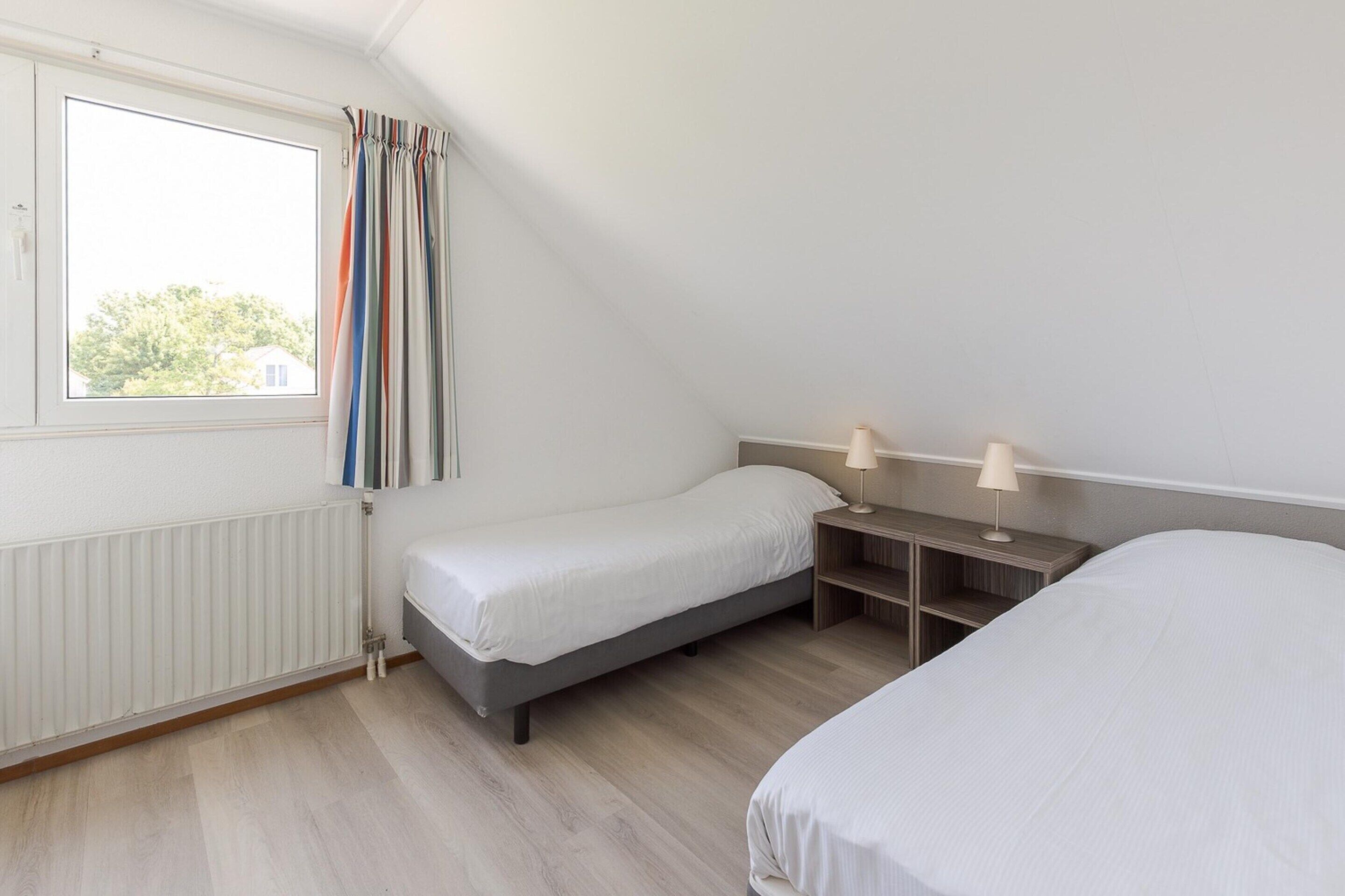 Un dormitorio con una cama individual, una mesita de noche con lámpara, una ventana con cortinas y un radiador.