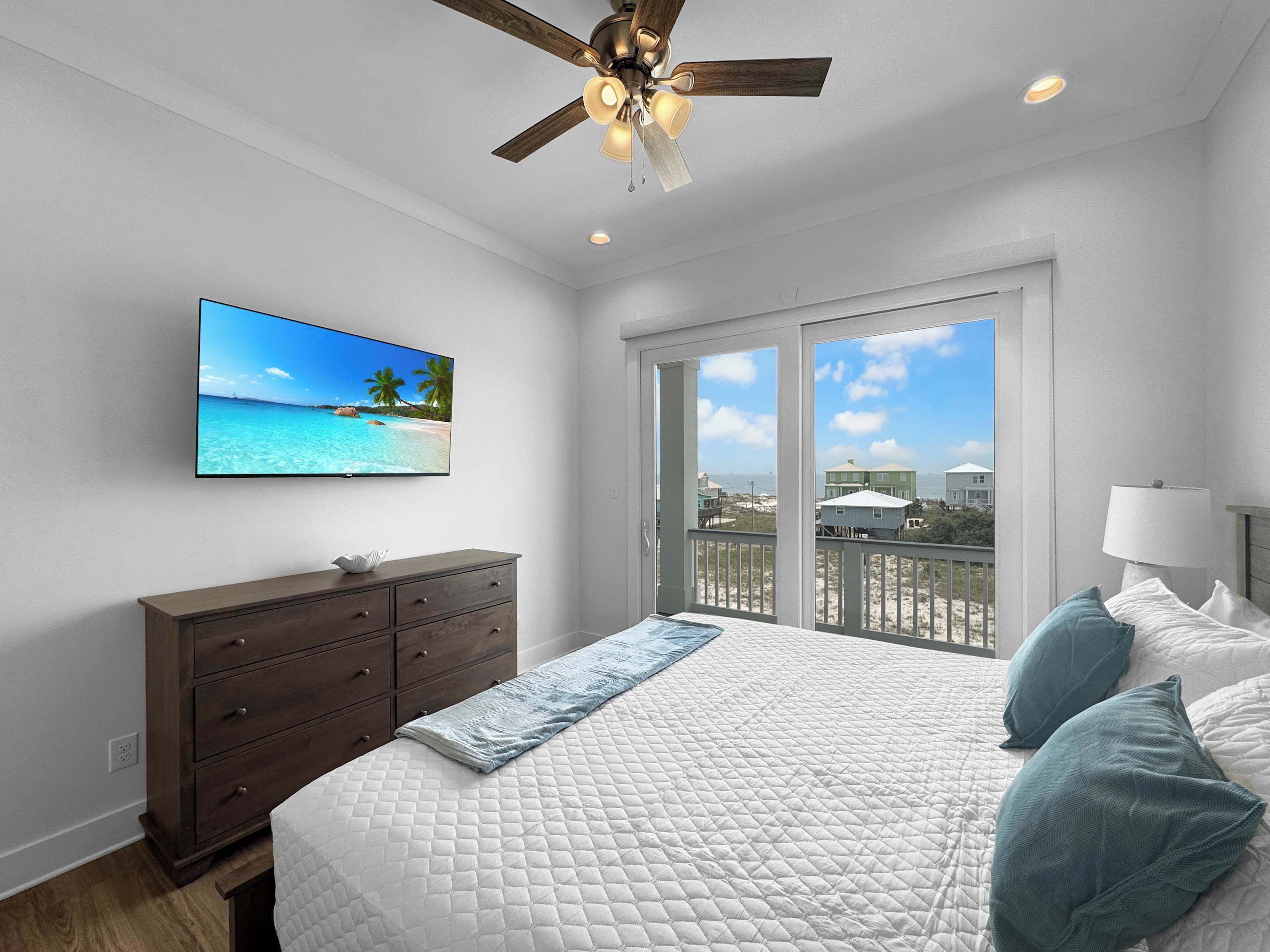 Un dormitorio con cama, cómoda, ventilador de techo, televisor que muestra una escena de playa y balcón con vista a la playa y casas.