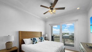 Quarto com cama de madeira, roupa de cama branca, ventoinha no teto e vista para a praia através de grandes janelas.