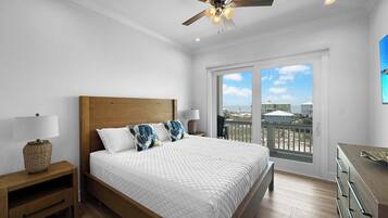 Un dormitorio con cama de madera, ropa de cama blanca, ventilador de techo y vista a la playa a través de amplios ventanales.