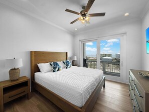 Un dormitorio con cama de madera, ropa de cama blanca, ventilador de techo y vista a la playa a través de amplios ventanales.
