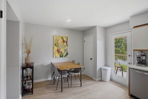 Een eetruimte met een tafel, stoelen, een schilderij aan de muur en uitzicht op het buitenleven door een deur.