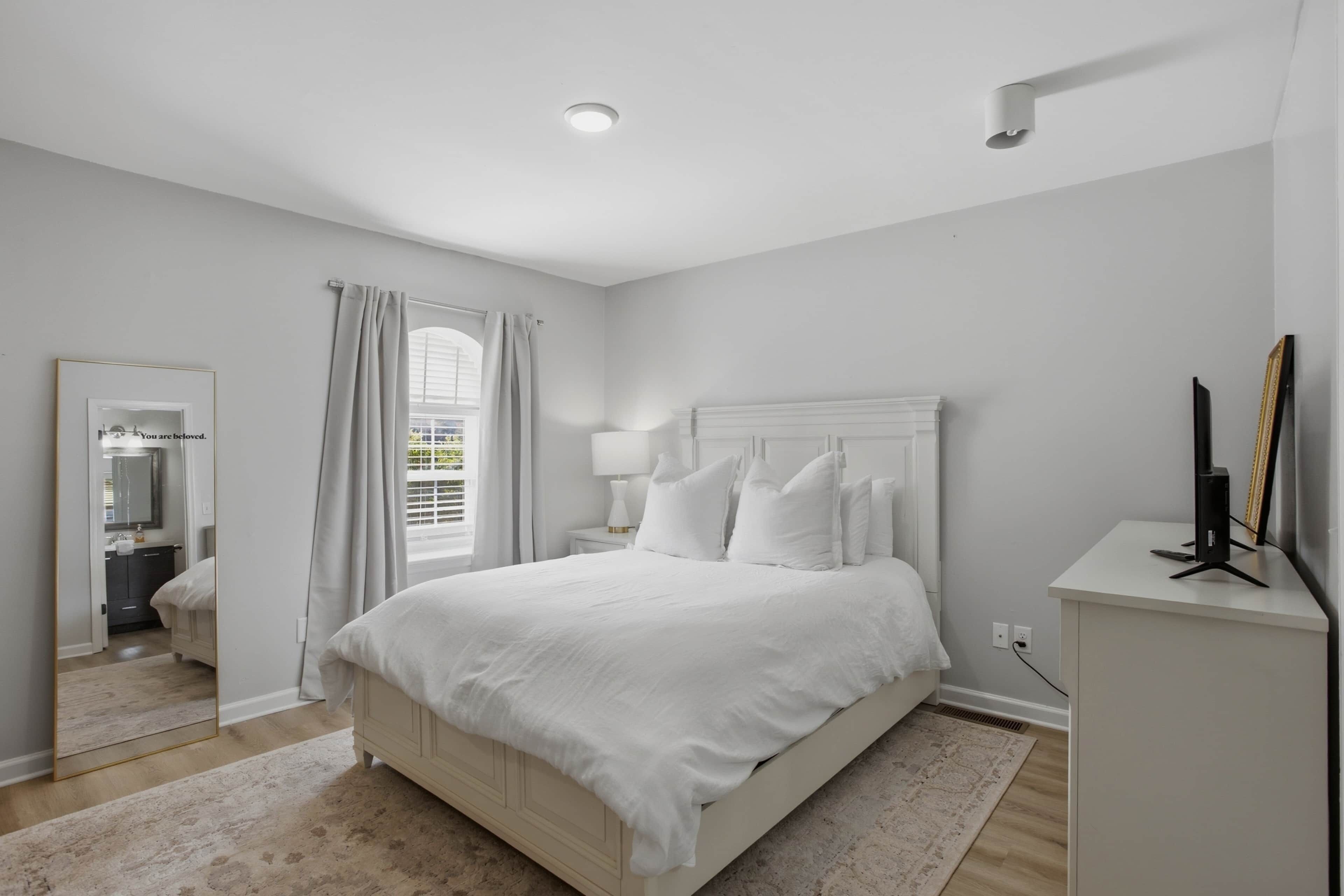 Un dormitorio con cama, mesita de noche, lámpara, espejo y ventana con cortinas.