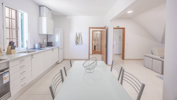 A modern kitchen with white cabinets, a dining table with chairs, and a view into the adjacent living area. - Tony's House B by YOUR KEY (Santana)