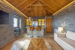A rustic interior with a stone wall, wooden ceiling, a bar area with stools, a sofa, and a lamp.