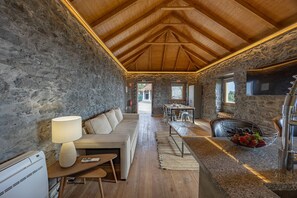 A room with a stone wall, wooden ceiling, a sofa, a table with a lamp, and a dining area with chairs.