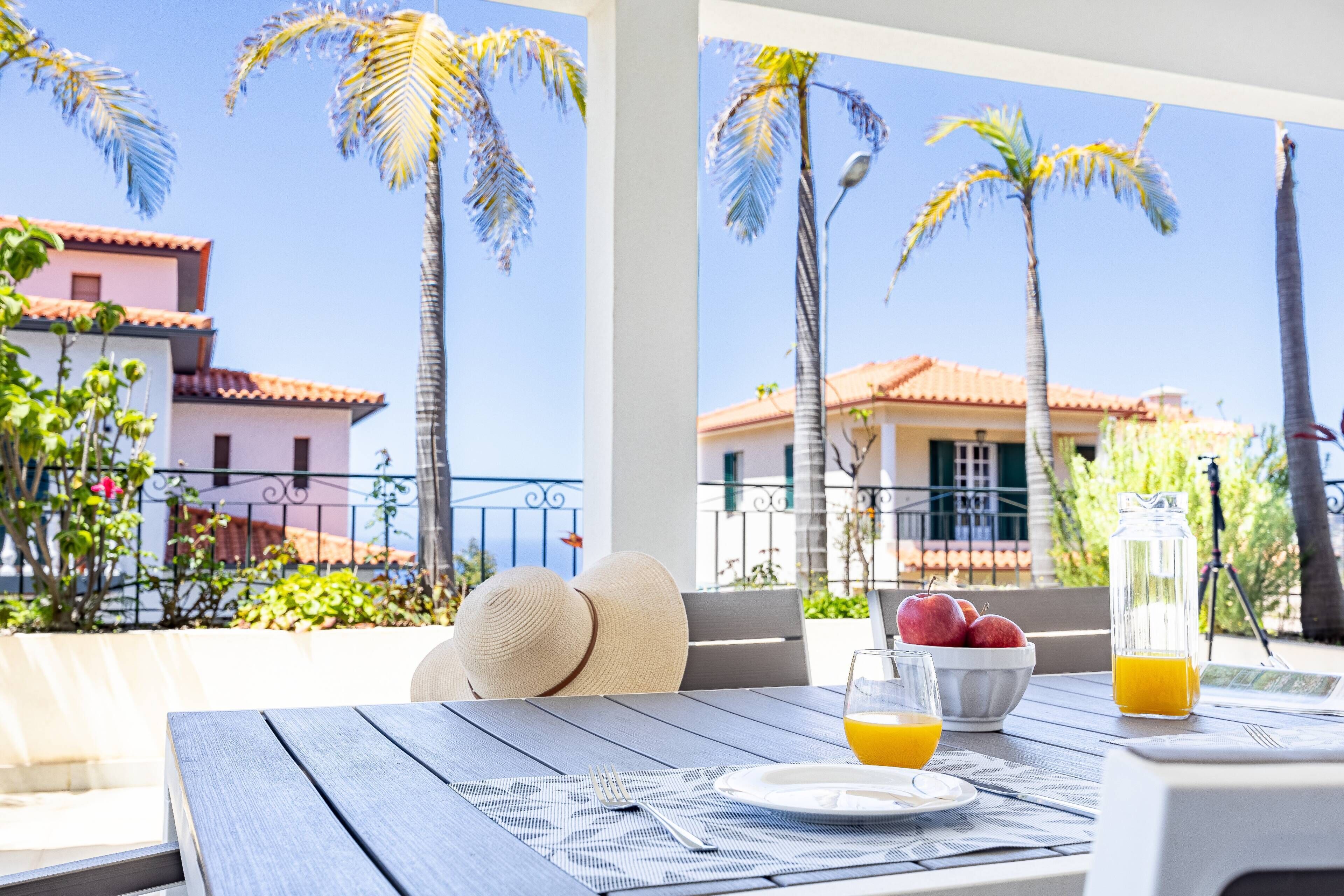 A table set with a plate, a bowl of fruit, and glasses of orange juice, with a view of palm trees and buildings in the background.