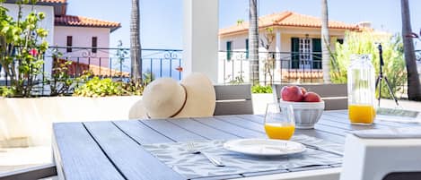 A table set with a plate, a bowl of fruit, and glasses of orange juice, with a view of palm trees and buildings in the background.