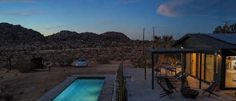 Una piscina con zona de patio, un coche y una casa en un paisaje desértico.