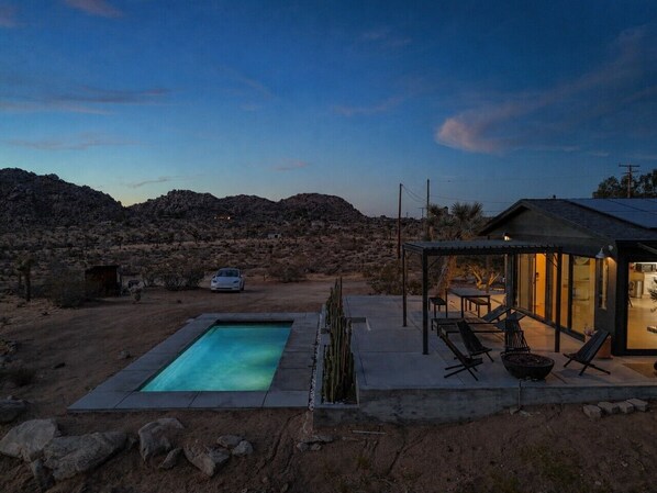 Una piscina con zona de patio, un coche y una casa en un paisaje desértico.