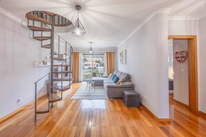 A modern living room with a wooden floor, a grey sofa, a staircase with a metal railing, and a view of houses through the window. - Vista Mar À Costa by YOUR KEY (Santa Cruz)