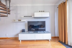 Un salon moderne avec un téléviseur à écran plat posé sur un meuble blanc, une petite table avec une télécommande et une étagère murale agrémentée d’objets décoratifs.
