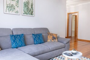Un canapé gris agrémenté de coussins à motifs, une petite table ronde sur laquelle reposent une télécommande et une plante, et deux reproductions botaniques encadrées accrochées au mur.