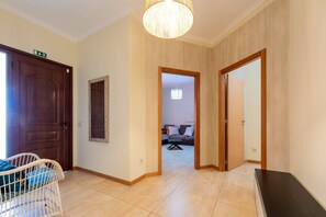 A hallway with a wooden door, a white wicker chair, a hanging light fixture, and a view into a living area with a sofa and a table. - Villa Areal I by YOUR KEY (Porto Santo)