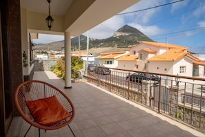 Un balcon avec un fauteuil en osier, un pilier en pierre et une vue sur des immeubles et des montagnes.