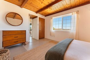 A bedroom with a wooden ceiling, a large window with curtains, a bed, a wooden dresser, and a mirror. - Bed N` Burke | Pool + Spa | Peloton | Fire Pit (Yucca Valley)