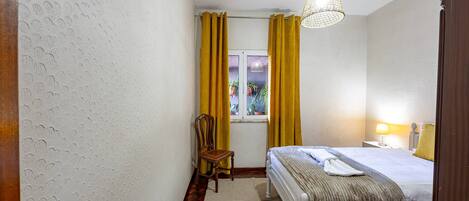 Habitación de hotel con cama, una silla, ventana con cortinas, lámpara colgante y lámpara de noche.