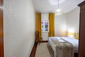 Une chambre d’hôtel avec un lit, une chaise, une fenêtre avec des rideaux, un luminaire suspendu et une lampe de chevet.