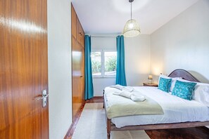 A bedroom with a wooden wardrobe, a bed with white bedding and blue pillows, a window with curtains, and a hanging light fixture. - Vivenda Paz Lobo by Your Key (Santa Cruz)