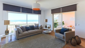 A modern living room with a sofa, armchairs, and a coffee table. Large windows offer a view of the beach.