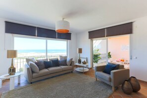 A modern living room with a sofa, armchairs, and a coffee table. Large windows offer a view of the beach.