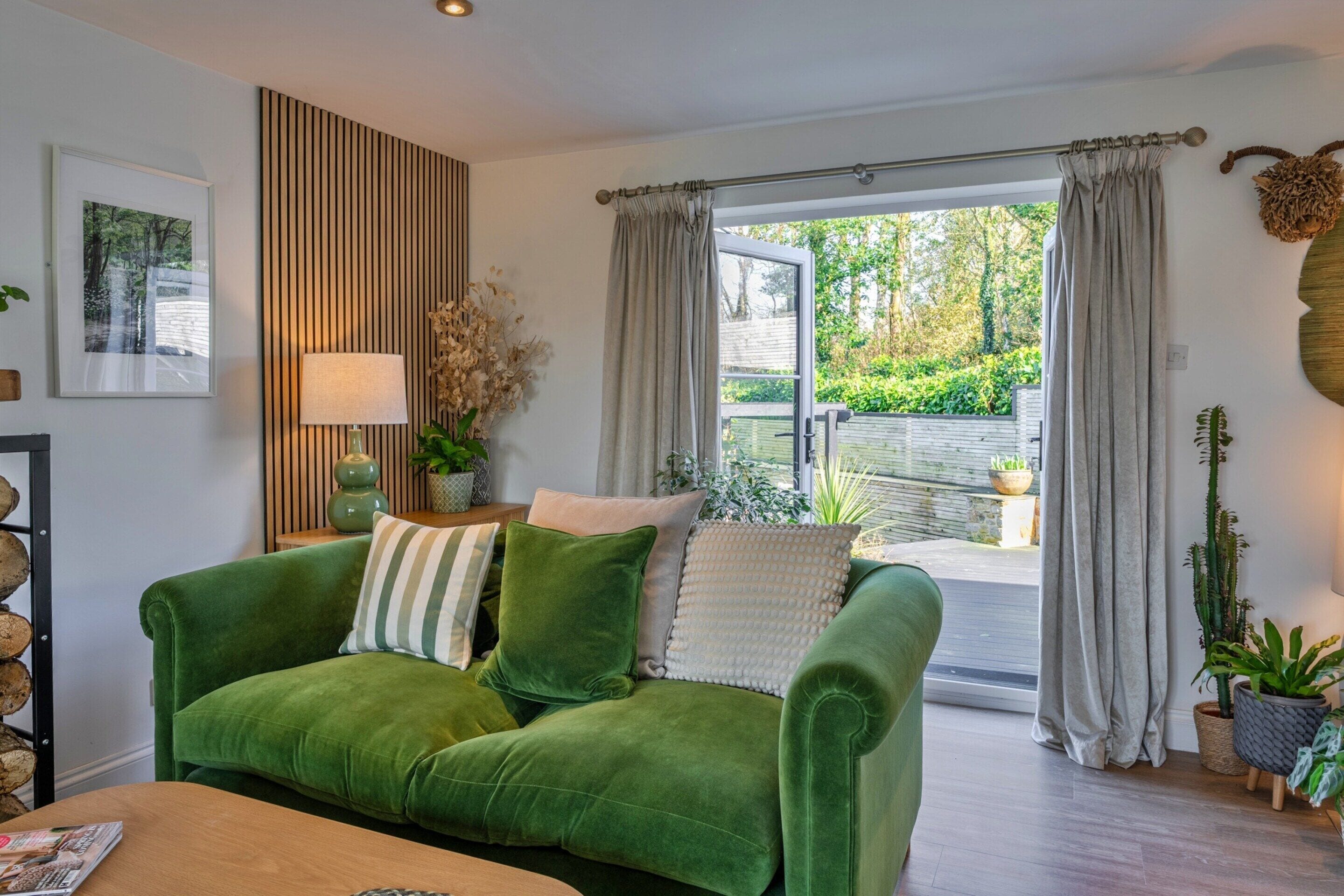 A green sofa with multiple cushions, a lamp, a framed picture, and a window with curtains.