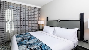 Quarto de hotel com cama, mesa de cabeceira, abajur e janela com cortinas.
