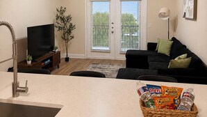 Een moderne woonkamer met een flatscreen televisie, een plafondventilator, een eethoek met een mand lekkernijen, en een glazen schuifdeur die leidt naar een terras.