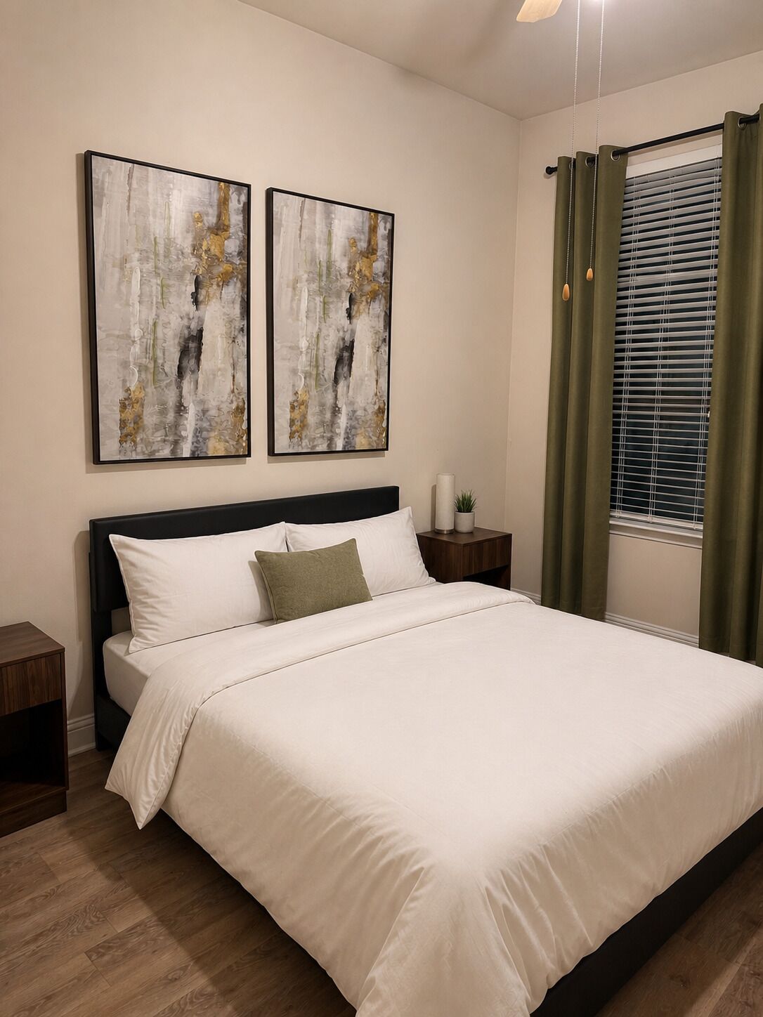 A neatly made bed with white bedding, two abstract paintings on the wall, a bedside table with a lamp, and a window with curtains.