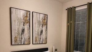 Een net opgemaakt bed met witte lakens, twee abstracte schilderijen aan de muur, een nachtkastje met een lamp, en een raam met gordijnen.