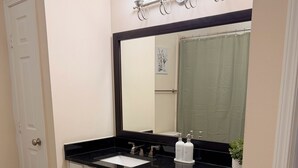 Een badkamer met een zwarte countertop, witte kastjes, een grote spiegel, een handdoekrek en een groene vloerkleed.