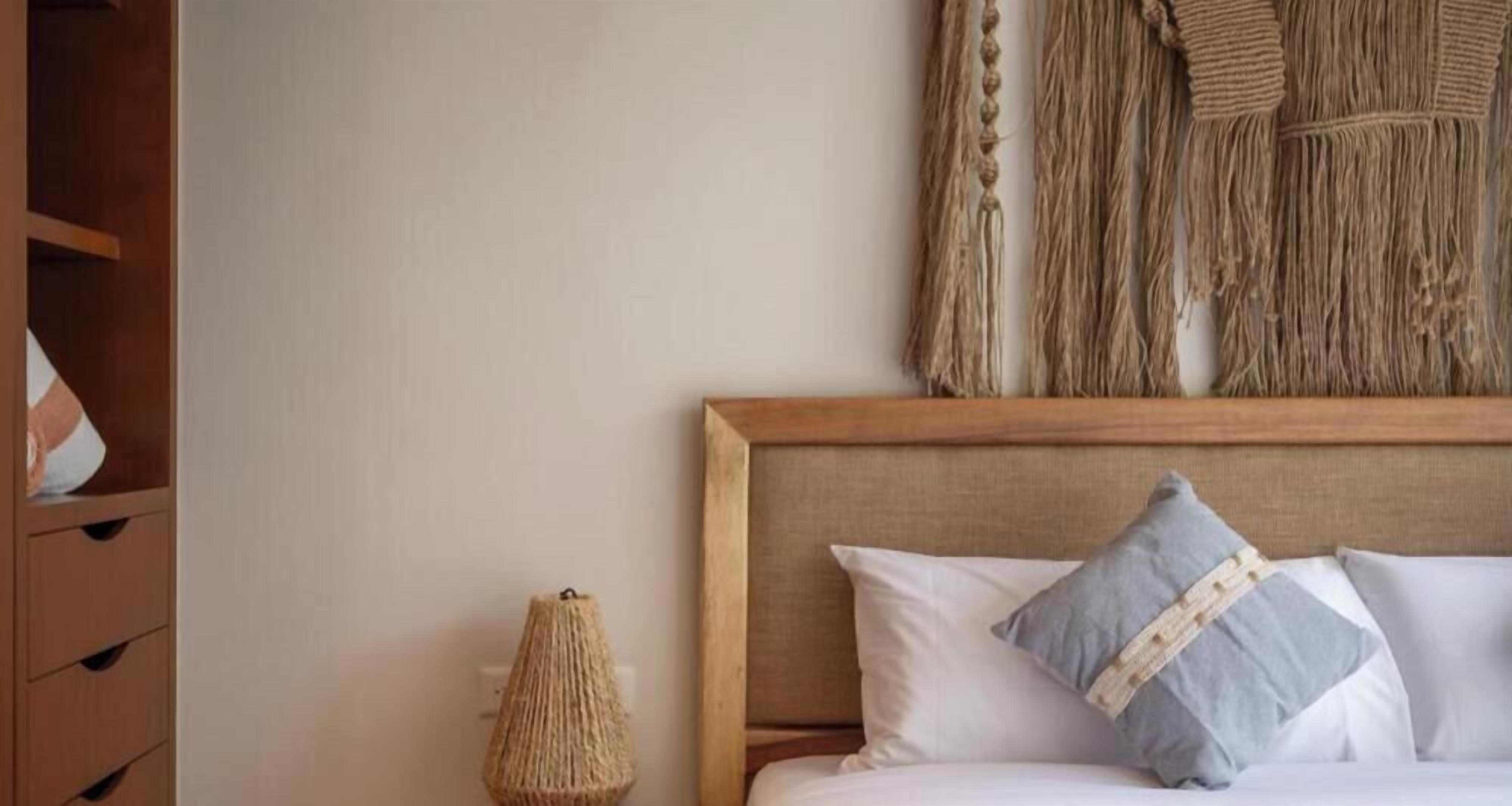 Un dormitorio con cabecera de madera, cama con almohadas blancas y grises, lámpara tejida y un tapiz de macramé en la pared.